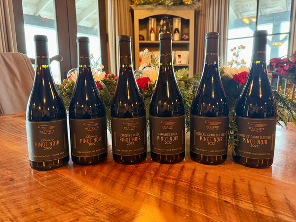 Six bottles of Westside Vineyards Pinot Noir on a wooden table with a blurred background. Vintages only available to wine club members.