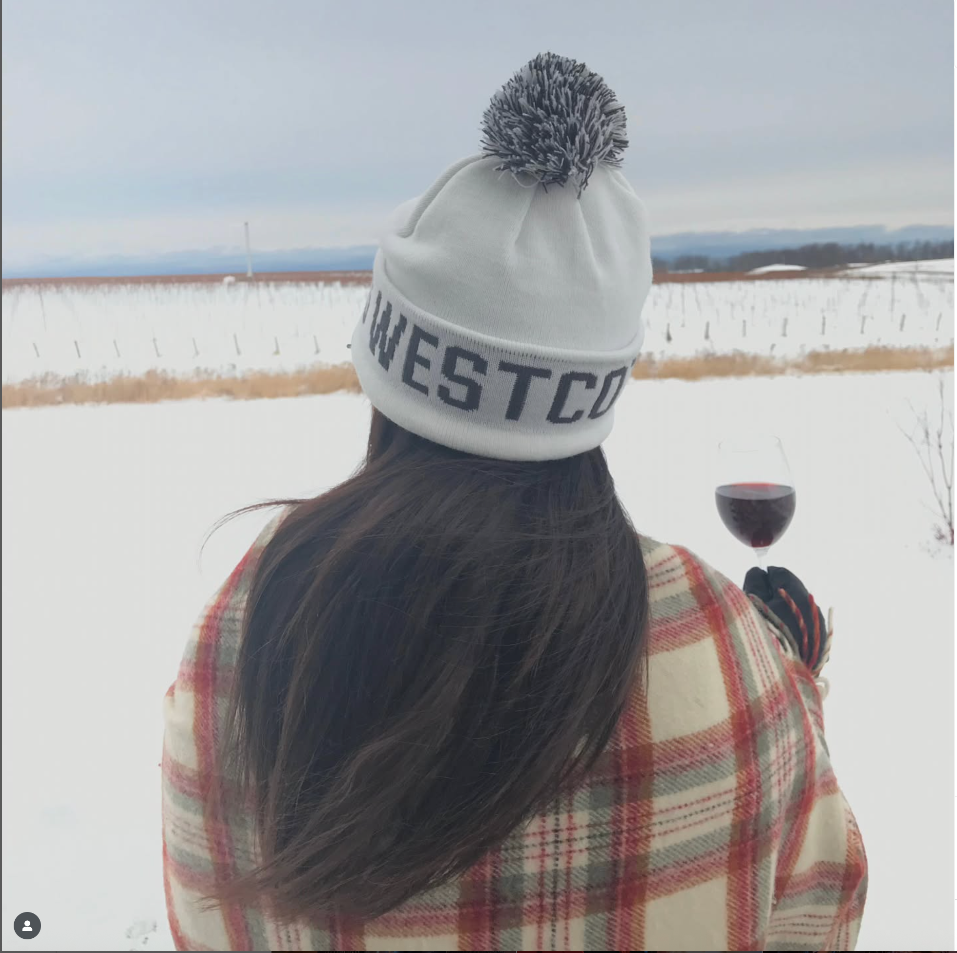 A woman with long brown hair wears a Westcott Vineyards toque and holds a glass of Pinot Noir as she overlooks the vineyard in winter. 